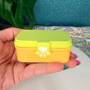 May include: A small, rectangular yellow and green box with a paw print clasp. The box has a bright yellow base and a lime green lid. The clasp is a paw print shape with a white heart in the center. The box is held in a hand.