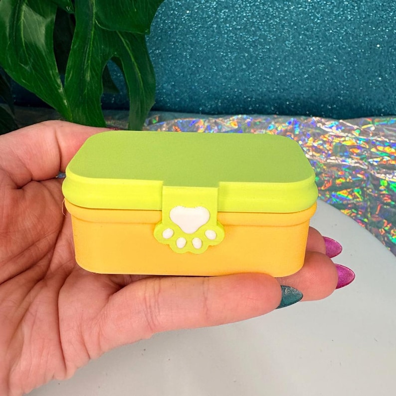 May include: A small, rectangular yellow and green box with a paw print clasp. The box has a bright yellow base and a lime green lid. The clasp is a paw print shape with a white heart in the center. The box is held in a hand.