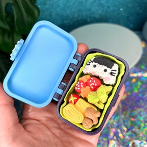 May include: A miniature blue bento box, open to reveal a colorful assortment of food items. The box contains a rice ball with a cat face, red tomatoes, yellow omelet, and other food items. The box is held in a hand.