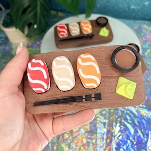Puede incluir: Un juego de sushi en miniatura con tres piezas de nigiri, con aderezos rojos, beige y naranjas, un pequeño plato de salsa marrón y un cuadrado de wasabi en una bandeja de madera. También se incluyen palillos negros.