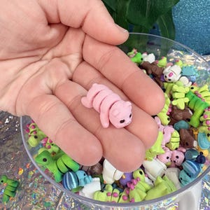 May include: A small, pink, segmented caterpillar figurine with black eyes and a smiling expression. The figurine is held in a hand, with a clear bowl filled with similar colorful figurines in the background. The figurines appear to be handmade.