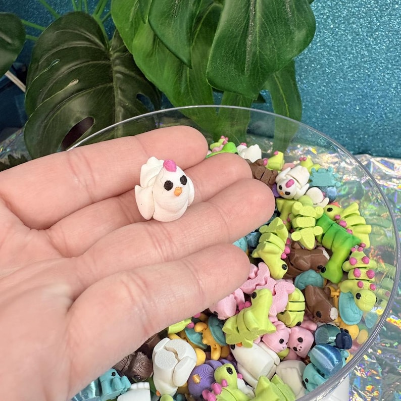 May include: A small, white polymer clay chick with a pink head crest and black eyes is held in a hand. The chick is surrounded by other colorful polymer clay figures in a clear bowl, including bunnies, dinosaurs, and Christmas trees.