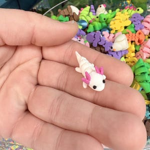 May include: A miniature white axolotl figurine with pink gills and black eyes. The small sculpture is held in a hand, with a blurred background of colorful polymer clay shapes.