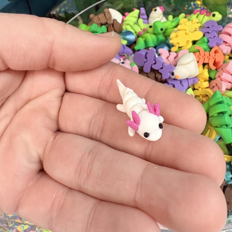 May include: A miniature white axolotl figurine with pink gills and black eyes. The small sculpture is held in a hand, with a blurred background of colorful polymer clay shapes.