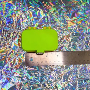 May include: A bright green rectangular plastic container with a hinged lid, measuring approximately 2.5 inches in length. The container is set against a shimmering, iridescent background. The container has yellow hinges and a white clasp.