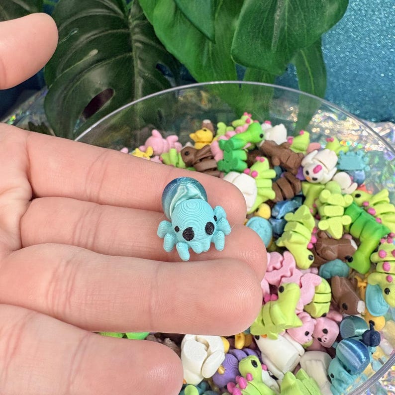 May include: A small, light blue octopus figurine with black eyes and a textured design. The octopus is held in a hand, with a clear bowl filled with colorful polymer clay charms in the background.
