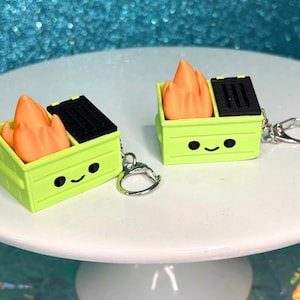 May include: Two keychains featuring a lime green dumpster with a black lid and orange flames. The dumpsters have a smiling face design. The keychains are on a white surface with a blue glitter background.