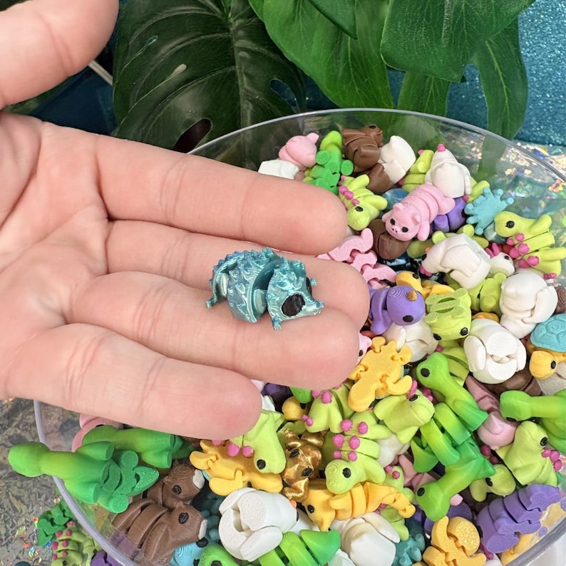 May include: A small, teal-colored hedgehog figurine with black eyes is held in a hand. The figurine is surrounded by a variety of colorful, miniature animal-shaped polymer clay pieces, including turtles, caterpillars, and other creatures.