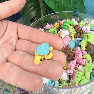 May include: A small, yellow turtle figurine with a light blue shell is held in a hand. The turtle has black eyes and is surrounded by other miniature animal figurines in a clear bowl. The bowl contains various colorful animal shapes.