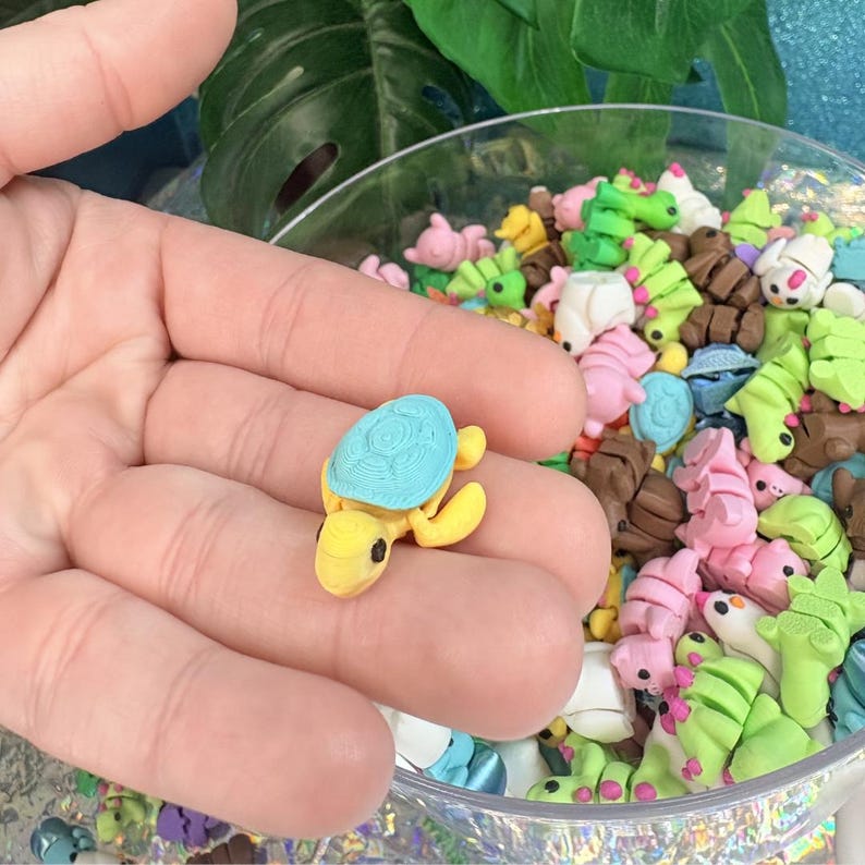 May include: A small, yellow turtle figurine with a light blue shell is held in a hand. The turtle has black eyes and is surrounded by other miniature animal figurines in a clear bowl. The bowl contains various colorful animal shapes.