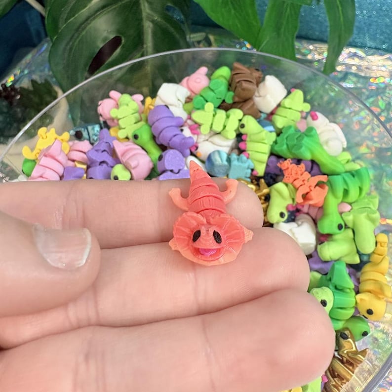 May include: A close-up of a small, coral-colored lizard-like polymer clay figure with a smiling face, held in a hand. The figure is surrounded by a variety of colorful polymer clay shapes in a clear bowl, including green, purple, and yellow.