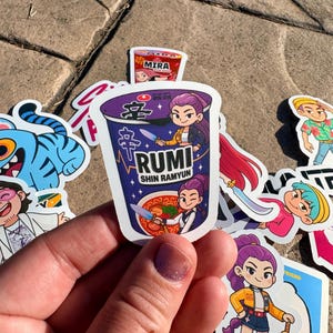 May include: A collection of colorful stickers featuring cartoon characters and food packaging. The central sticker is a purple ramen cup with the text "RUMI SHIN RAMYUN" and an illustration of a woman holding a knife. Other stickers include a blue tiger and a small "MIRA" ramen cup.