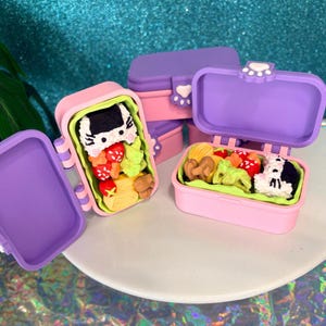 May include: Miniature pink and purple bento boxes, some open to reveal detailed food replicas. One box features a cat-shaped rice ball, while another has a dog-shaped rice ball. The boxes have paw-shaped clasps.