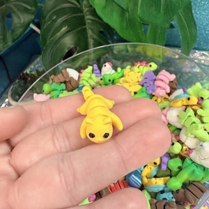 May include: A small, yellow polymer clay cat figurine with black eyes and a detailed body. The cat is lying down, surrounded by other colorful clay animal figurines in a clear bowl. The background includes green leaves and a blue surface.