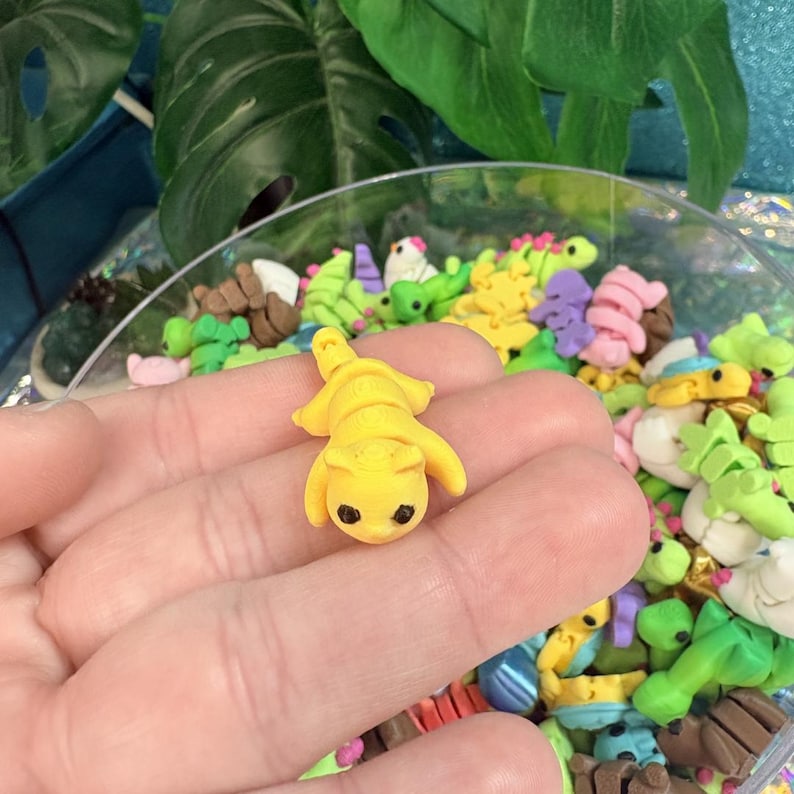 May include: A small, yellow polymer clay cat figurine with black eyes and a detailed body. The cat is lying down, surrounded by other colorful clay animal figurines in a clear bowl. The background includes green leaves and a blue surface.