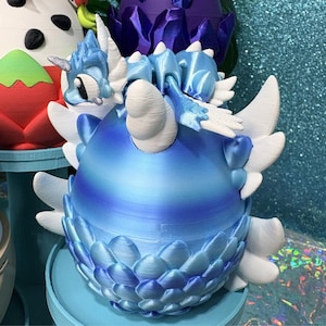May include: A 3D printed blue and white dragon figurine emerging from a matching blue egg. The dragon features large white wings, a detailed head with a horn, and a scaly texture on the egg base. The entire piece displays a gradient of blue.