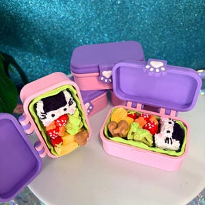 May include: Pink and purple miniature bento boxes with cat paw closures. The open boxes reveal detailed food replicas, including sushi, vegetables, and fruit. The bento boxes are stacked on a light blue surface.