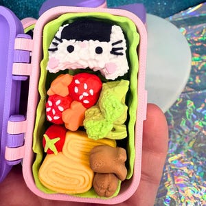 May include: A miniature, open lunchbox with a variety of food items. The lunchbox is pink and purple, with a green interior. The food includes a cat-shaped rice ball, strawberries, and other colorful items. The lunchbox is held in a hand.