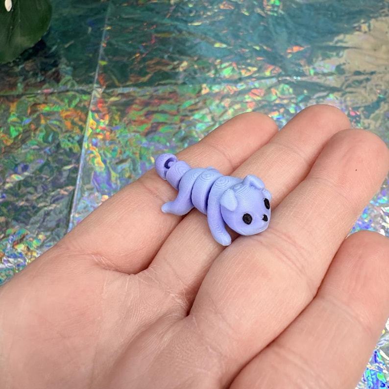 May include: A small, lavender-colored figurine of a stylized animal with a curled tail and black eyes. The figurine is lying on a surface. The animal has a segmented body and a rounded head with small ears.