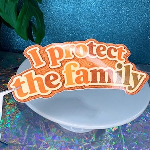 May include: A sticker with the text "I protect the family" in orange and brown lettering. The sticker has a wood grain background and is placed on a light blue surface. The background is a sparkly blue.