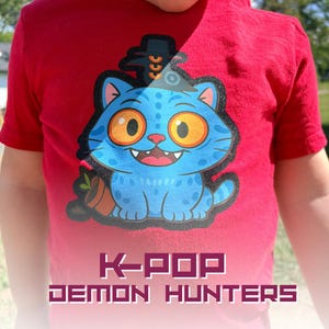 May include: Red t-shirt featuring a blue cartoon cat with orange eyes, a black hat, and a small pot with a plant. The text "K-POP DEMON HUNTERS" is printed at the bottom of the shirt. The shirt is made of a soft material.