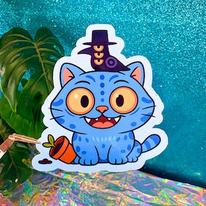 Puede incluir: Una pegatina de gato azul con grandes ojos naranjas, un cuervo con sombrero y una maceta volcada. El gato tiene una sonrisa traviesa y está cubierto de pequeños puntos azules. La pegatina está sobre un fondo de purpurina verde azulado.