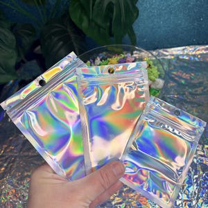 May include: Three iridescent, holographic zip-top bags are held in a hand. The bags reflect a rainbow of colors, including pink, green, and blue. Each bag has a small hole near the top for hanging. The background is a mix of plants and a glittery surface.