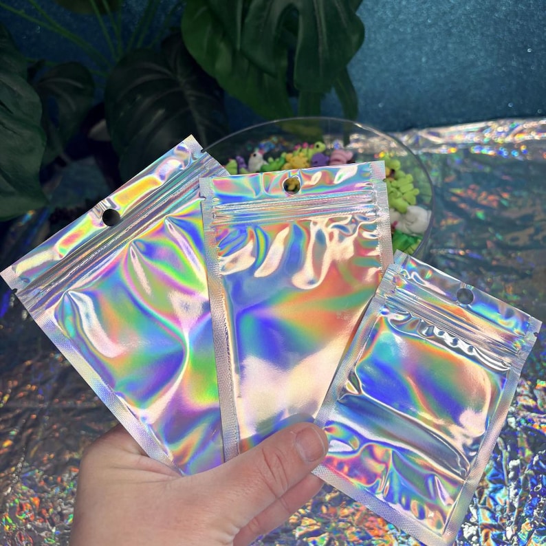 May include: Three iridescent, holographic zip-top bags are held in a hand. The bags reflect a rainbow of colors, including pink, green, and blue. Each bag has a small hole near the top for hanging. The background is a mix of plants and a glittery surface.