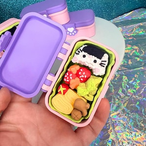 May include: A miniature, open bento box with a purple lid and pink base, filled with colorful food replicas. The food includes sushi, strawberries, and other items. Additional closed bento boxes are visible in the background. The bento box is held in a hand.