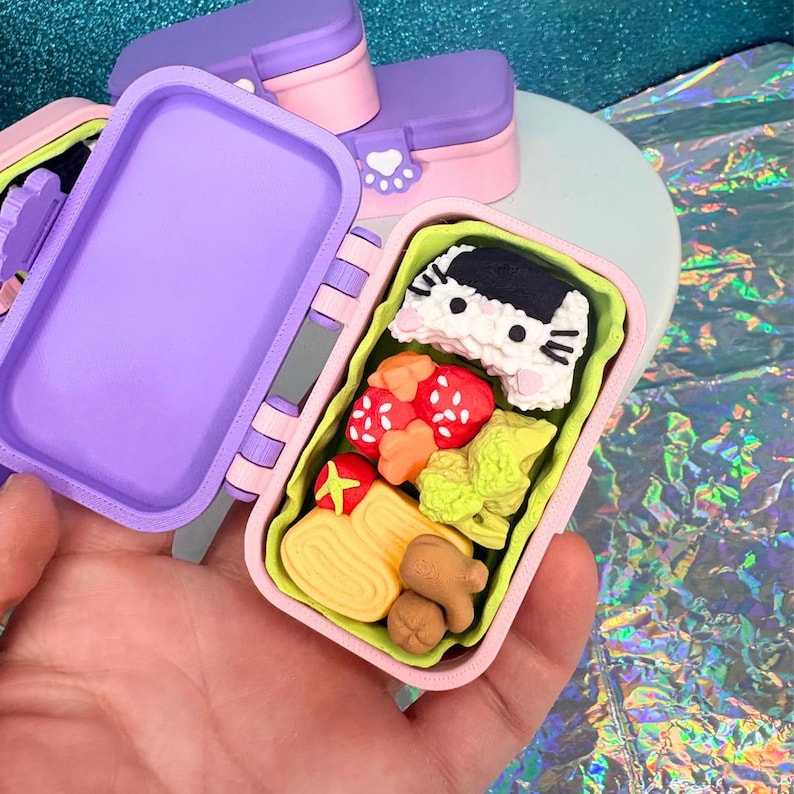 May include: A miniature, open bento box with a purple lid and pink base, filled with colorful food replicas. The food includes sushi, strawberries, and other items. Additional closed bento boxes are visible in the background. The bento box is held in a hand.
