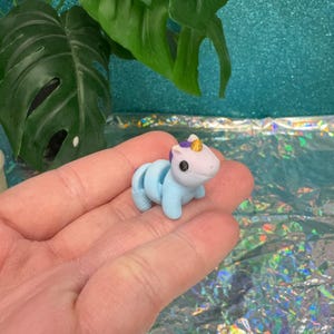 May include: A small, light blue unicorn figurine with a white face, purple mane, and gold horn. The toy is held in a hand, with a green leafy plant and a shimmering background visible.