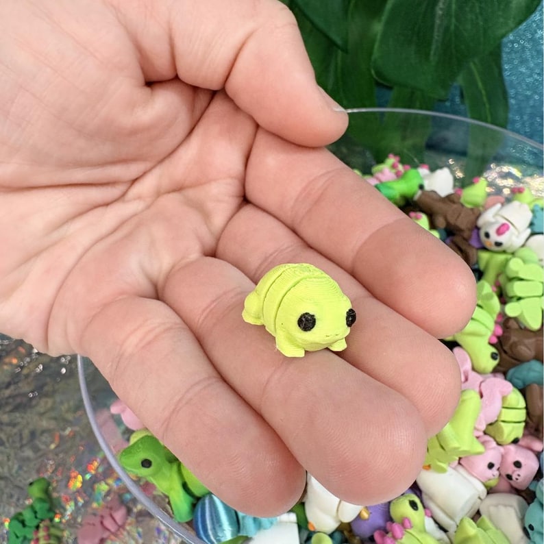 May include: A small, lime green turtle figurine with black eyes rests in an open hand. The turtle is segmented and has a smooth texture. Other colorful miniature animal figurines are visible in the background.