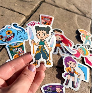May include: A collection of colorful stickers featuring cartoon characters. One sticker depicts a girl with two swords. Other stickers include a blue tiger, a girl with red hair, and a girl with purple hair.