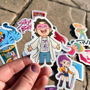 May include: A collection of colorful stickers featuring cartoon characters and text. The central sticker depicts a smiling character wearing a pink eye mask, white coat, and blue jeans. Other stickers include a blue tiger, a character with purple hair, and text that reads "ZOEY".