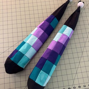 Peut inclure: Deux longs tubes en tissu effilés présentant un motif en patchwork de carrés alternés violets, bleu sarcelle et bleu clair. Chaque tube possède une base en tissu noir uni et une section supérieure noire terminée par un petit pompon blanc.