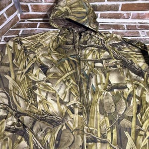 May include: A hooded camouflage jacket in shades of green, brown, and beige, designed to blend with natural environments. The jacket has a zip closure and a pocket. The background is a brick wall.