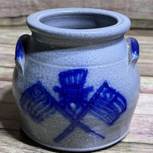 May include: A blue and gray ceramic jar with two handles. The jar features a blue design of crossed flags and a top hat. The jar is a decorative piece.