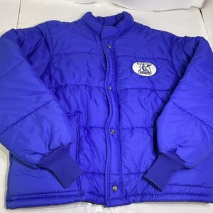 May include: A vibrant blue puffer jacket with a quilted design. The jacket features a stand-up collar, snap closures, and ribbed cuffs. A circular patch with a graphic is on the upper left chest. This outerwear is suitable for cold weather.