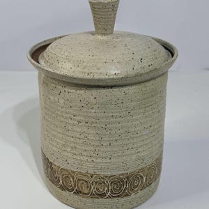 May include: A beige ceramic storage jar with a lid and a handle. The cylindrical jar features a decorative band of brown spiral designs. The lid fits securely, and the handle is a small, cone-shaped knob. The jar is approximately 20 cm tall.