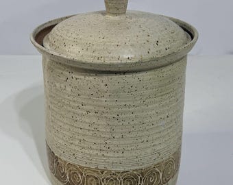 May include: A beige ceramic canister with a lid and a handle. The canister has a cylindrical shape with a decorative band of brown spiral designs around the base. The lid fits snugly on top, and the handle is a small, cone-shaped knob.