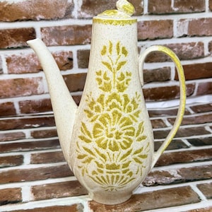 May include: Cream-colored ceramic coffee pot with a long spout and handle. The pot features a yellow floral design and a matching lid. The handle and rim are also yellow. The pot is set against a brick background.