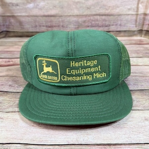 May include: Green John Deere Heritage Equipment Chesaning Mich cap with a mesh back and a curved peak. The front has a rectangular patch with the John Deere logo and text.