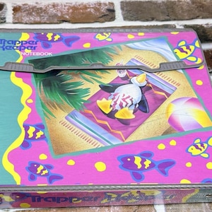 May include: A Trapper Keeper notebook featuring a pink and teal background with yellow fish patterns. The center graphic depicts a cartoon penguin wearing sunglasses and a patterned swimsuit, lounging on a beach towel under palm trees with a beach ball.