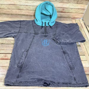 May include: A grey hooded pullover with a turquoise hood and a quarter-zip. The pullover features a light blue emblem on the chest and two front pockets. The garment has long sleeves and a drawstring at the bottom.