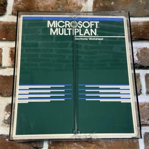 May include: A vintage Microsoft Multiplan Electronic Worksheet software package encased in a clear plastic protective box. The cover features a dark green background with white text, a blue horizontal bar, and a series of thin blue and white lines.