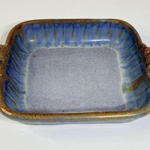 May include: A square ceramic serving dish with a blue and brown glaze. The interior is a speckled light blue, while the rim and handles are brown. The handles are decorative, with a knot-like design. A unique piece for serving food.