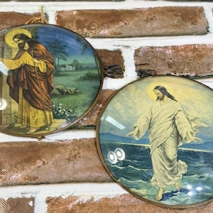 May include: Two circular wall plaques featuring religious imagery. The left plaque depicts a figure in a yellow and brown robe standing by a doorway. The right plaque shows a figure in a white robe walking on water against a blue sea background.