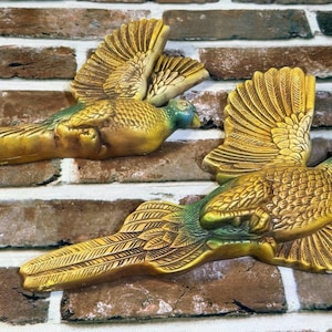 May include: Two wall-mounted bird sculptures featuring golden-yellow plumage with green accents on the necks and heads. Each bird is depicted in a flying position with wings spread wide and long, detailed tail feathers extending horizontally.