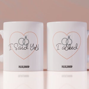 Puede incluir: Dos tazas de cerámica blanca con texto negro e ilustraciones. La taza de la izquierda dice "¡Dije que sí!" con un corazón y dos anillos. La taza de la derecha dice "Pregunté" con un corazón y dos anillos. Ambas tazas tienen la fecha "13.12.2020" impresa en la parte inferior.