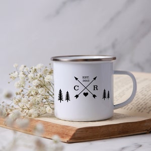 May include: White enamel camping mug with a black design featuring two pine trees, a heart, and the letters "C" and "R" with crossed arrows. The text "EST. 2012" is above the letters.